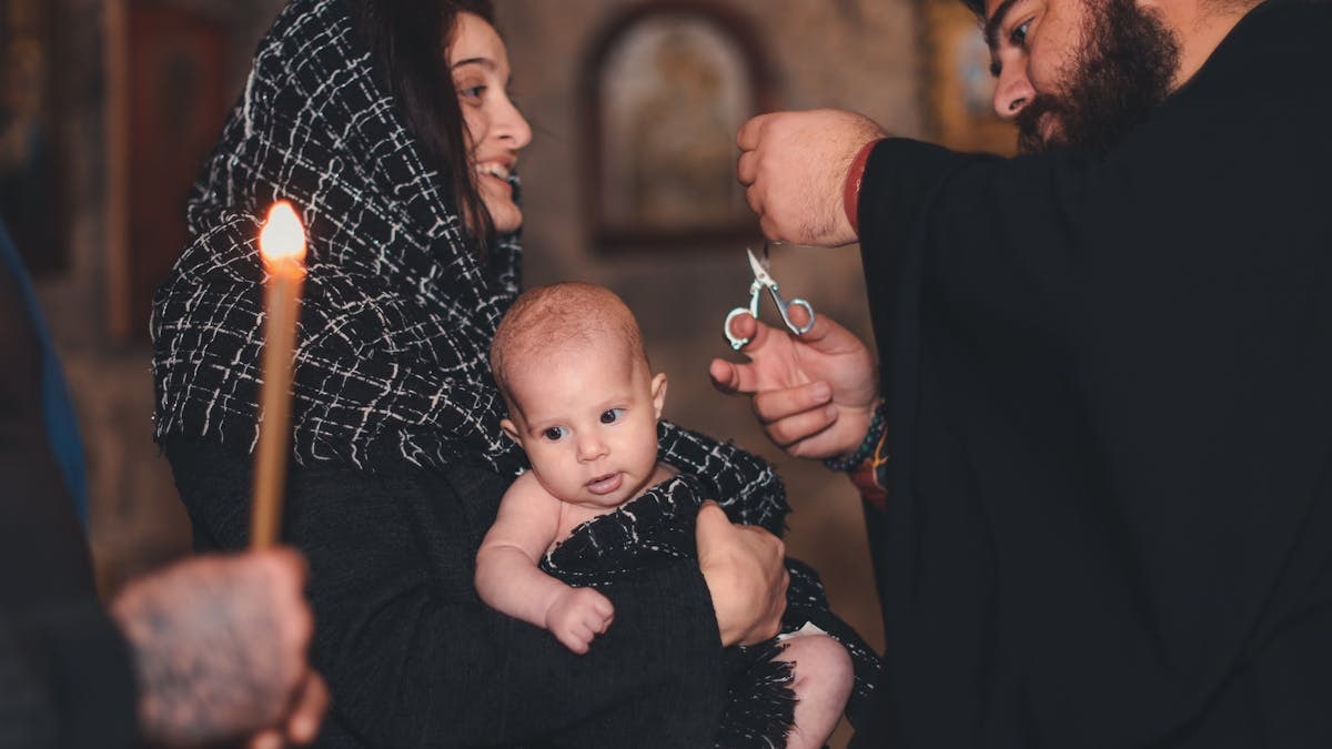 An intimate Orthodox baptism ceremony featuring a priest and family with candle.